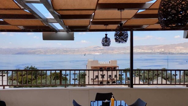 Een balkon met uitzicht op de zee en verre bergen, met een tafel, stoelen en een hanglamp.