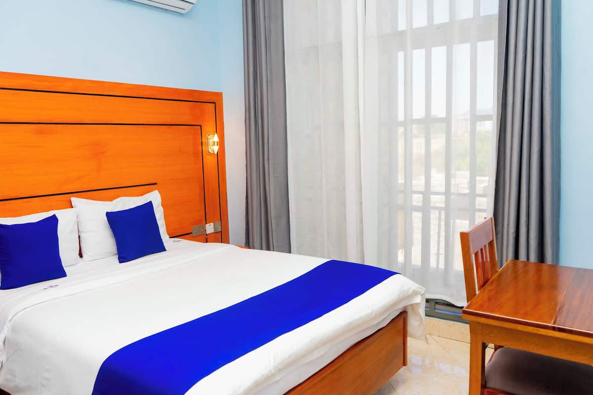 A hotel room with a wooden headboard, a bed with white and blue bedding, a wooden table, and a view through sheer curtains.