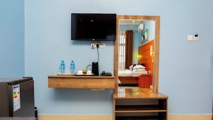 A hotel room with a TV mounted on the wall, a wooden shelf with toiletries, a bed with a headboard, and a mirror reflecting the room. - La Stanar Hotel (Dodoma)