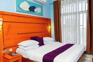 Una habitación de hotel con cabecera de madera, ropa de cama blanca, una manta morada y un ventanal con cortinas.