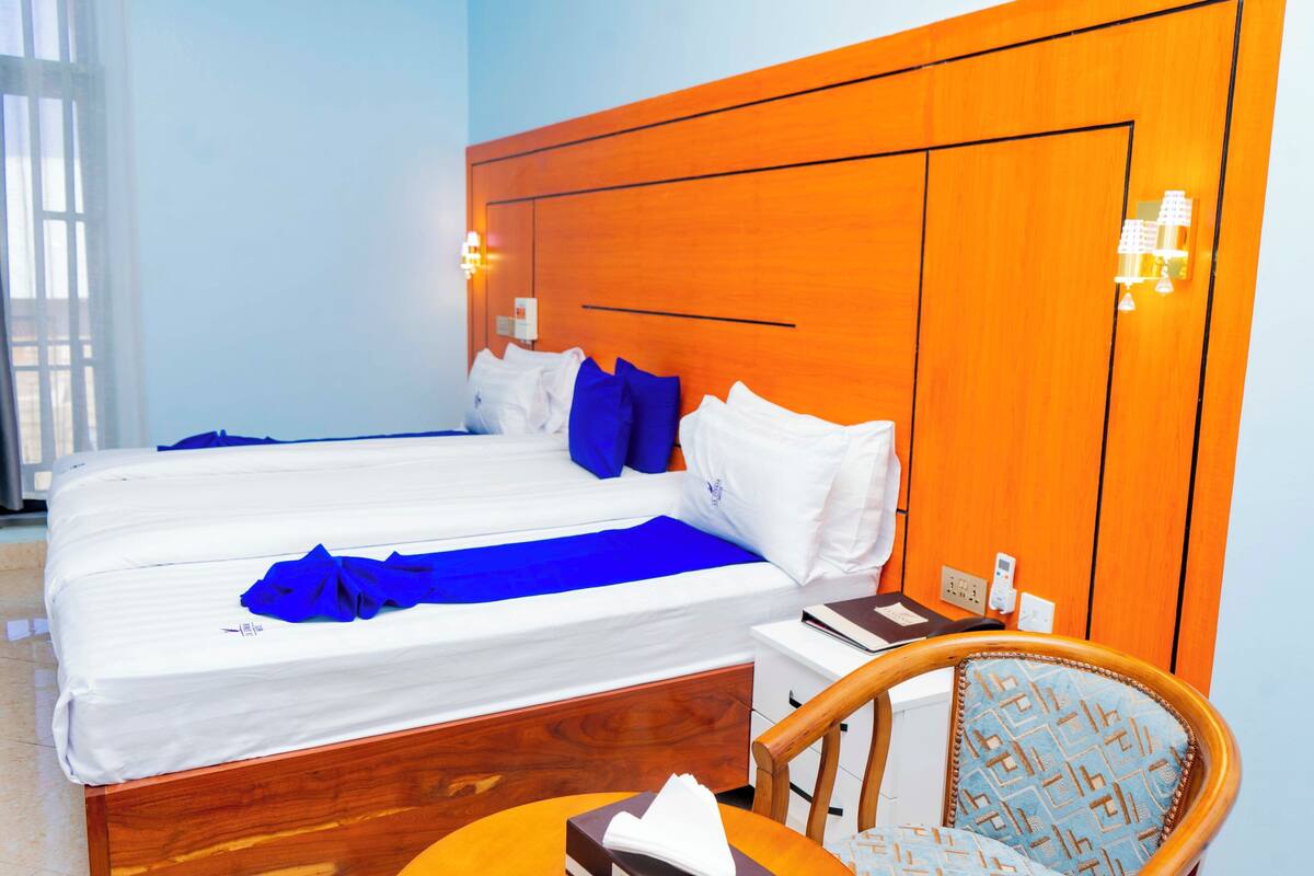 A hotel room with two beds, a wooden headboard, a bedside table with a lamp, and a chair with patterned upholstery.