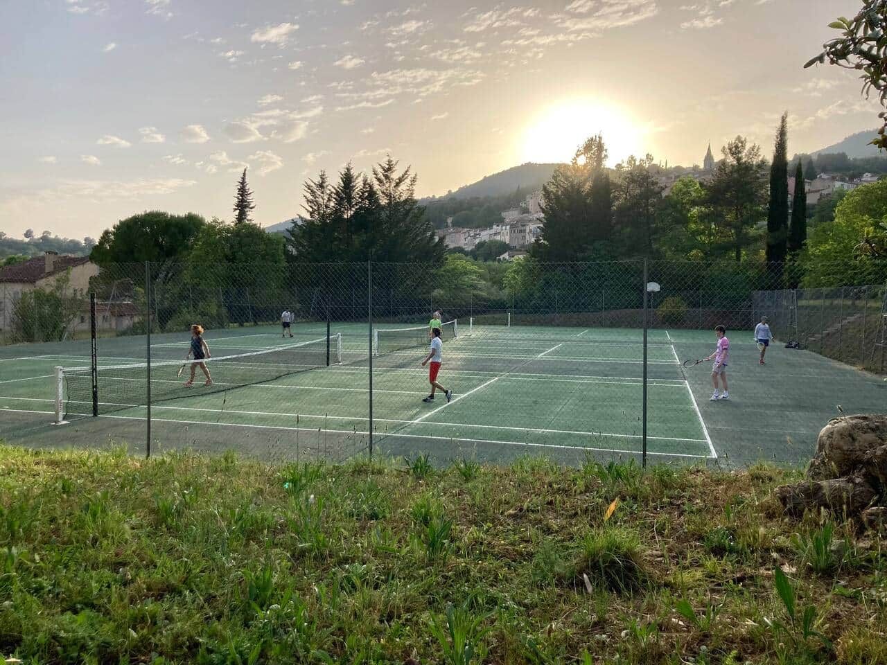 An outdoor tennis court with a net, surrounded by greenery and a fence.