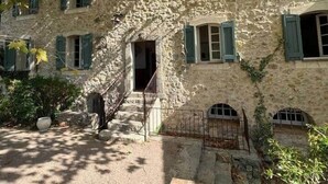 Un bâtiment en pierre doté d’un escalier central, de nombreuses fenêtres avec volets et d’un jardin qui l’entoure.