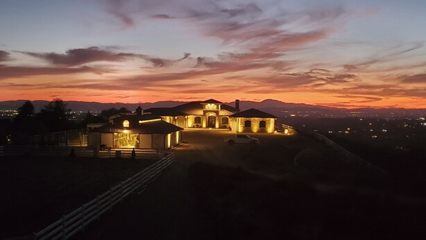 A large, illuminated building with multiple structures, including a main house and a smaller annex, set against a sunset sky. - Romantic 1 Bedroom Villa Overlooking the Vineyards with INCREDIBLE VIEWS! (Temecula)