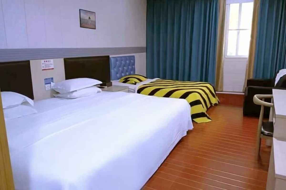 A hotel room with two beds, one with a striped bedspread and the other with a plain white bedspread. There is a wooden floor, a window with curtains, and a small desk with a chair.