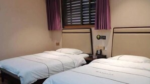 Une chambre d’hôtel avec deux lits simples, chacun doté d’un matelas blanc et d’oreillers assortis. Il y a une petite table de chevet avec une lampe, et une horloge est accrochée au mur au-dessus des lits.