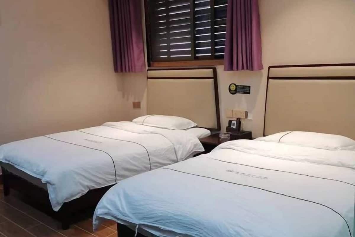 Habitación de hotel con dos camas individuales, cada una con un colchón blanco y almohadas a juego. Hay una mesita de noche pequeña y un reloj en la pared sobre las camas.
