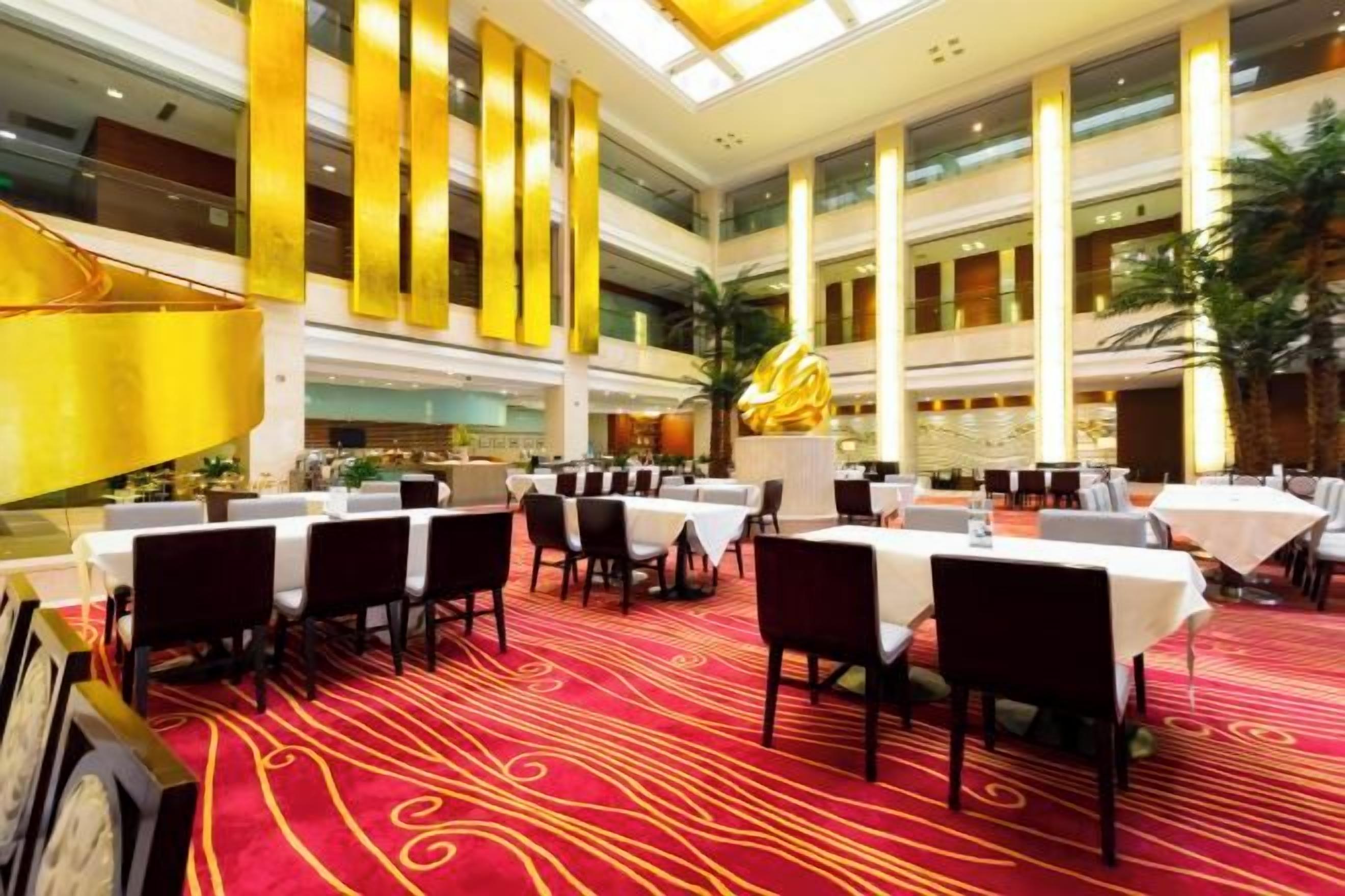 A spacious indoor dining area with multiple tables and chairs, high ceilings, and a central decorative sculpture.