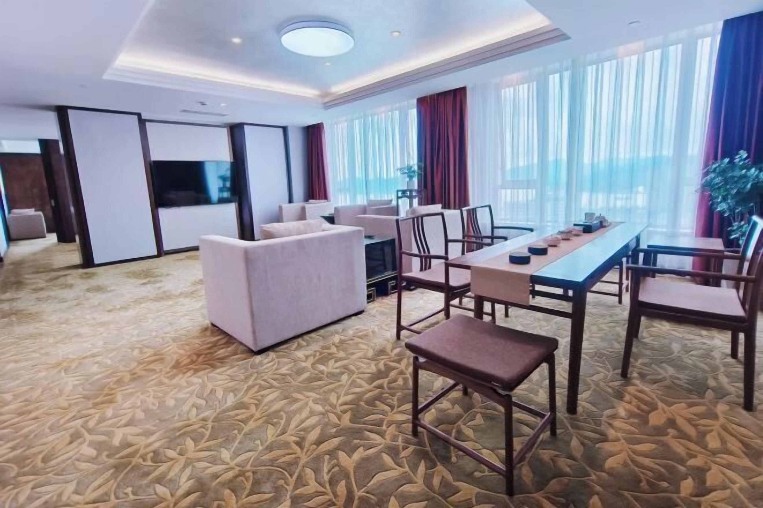 Quarto de hotel com uma cama grande, área de estar com sofá e cadeiras, mesa de jantar com cadeiras e uma janela ampla com cortinas.