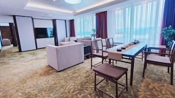 Quarto de hotel com uma cama grande, área de estar com sofá e cadeiras, mesa de jantar com cadeiras e uma janela ampla com cortinas.