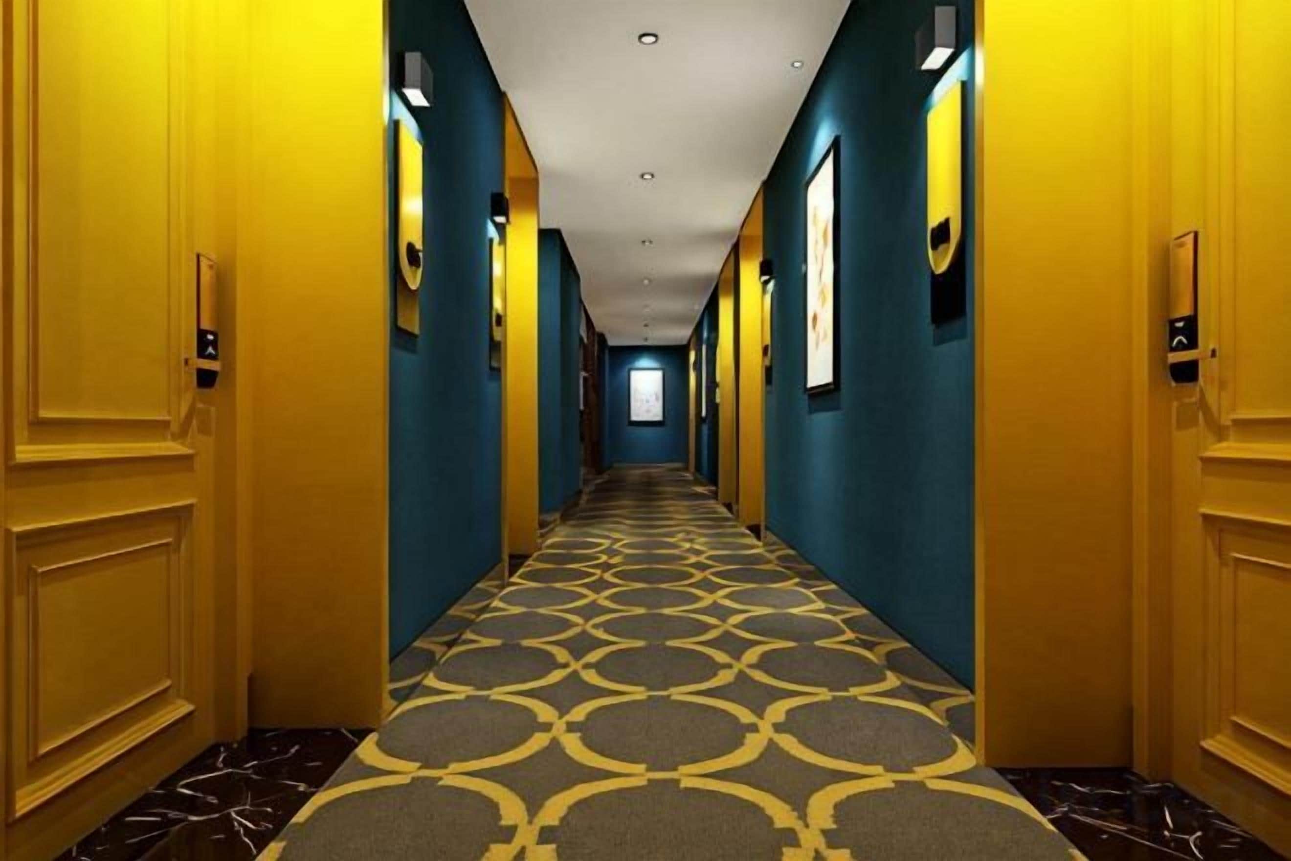 A hotel corridor with doors on both sides, a patterned carpet, and a ceiling with recessed lighting.