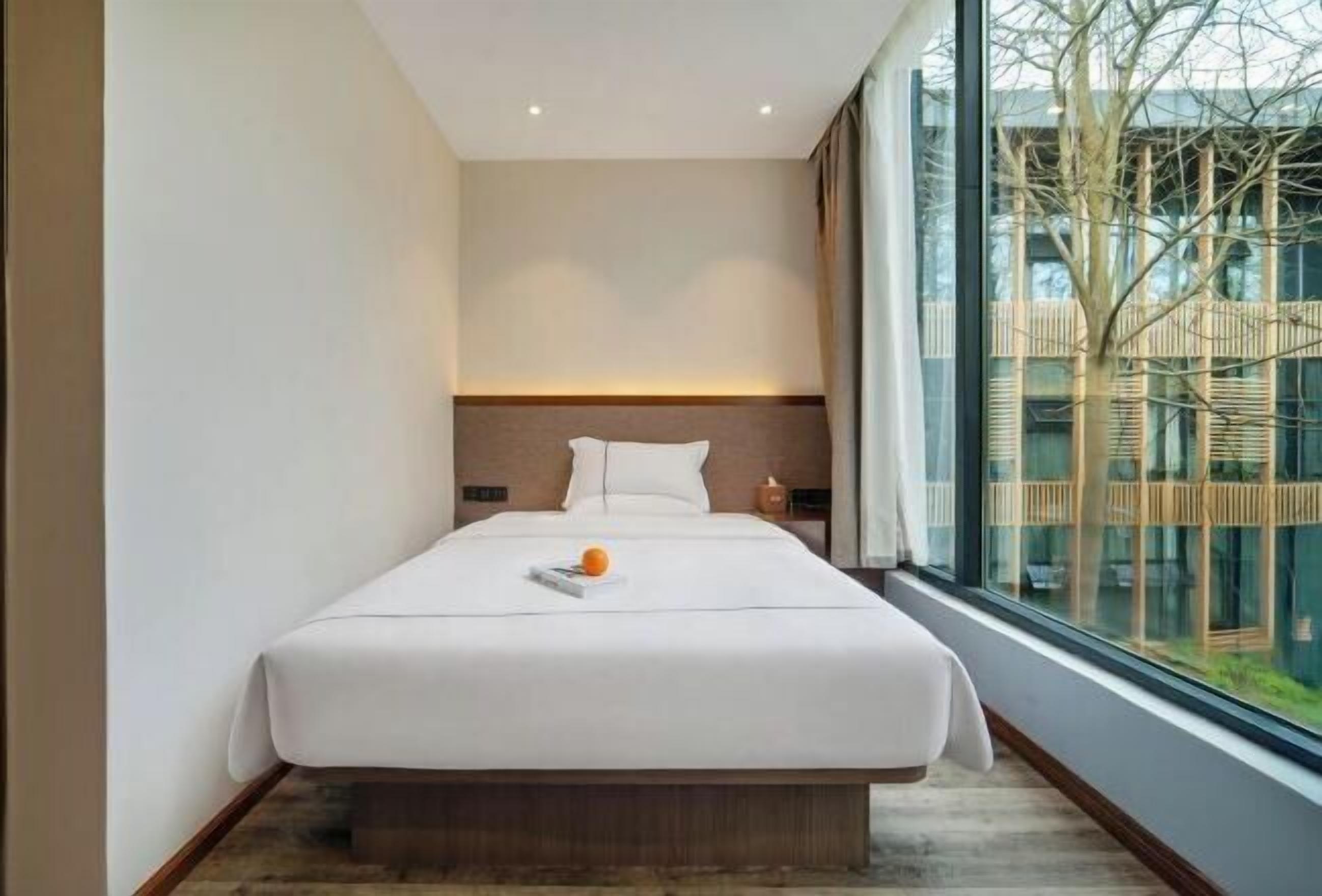 A modern hotel room with a large bed, a nightstand, and a view of a building through a large window.