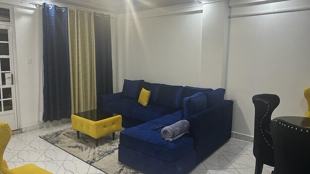 A living room with a blue sofa, yellow ottoman, and a glass coffee table. - Nanyuki One Bedroom Apartment (Nanyuki)