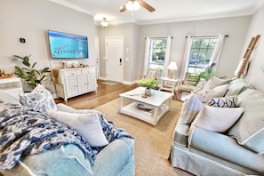 A living room with a television, white furniture, a coffee table, and a view of the outdoors.