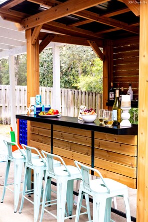 A covered outdoor bar area with wooden beams, a black countertop, and white bar stools.