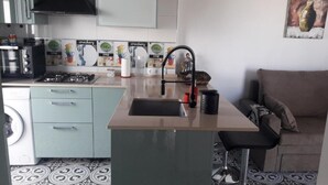 Cozinha compacta com pia, fogão e micro-ondas. Há uma área de jantar com uma cadeira e um sofá.