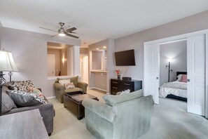 A modern living room with a sofa, armchair, and coffee table. There is a ceiling fan, a TV mounted on the wall, and a bed in the adjacent room.