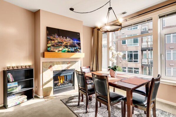 A dining area with a fireplace, a large window with a city view, and a bookshelf with books.