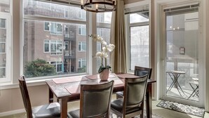 Un comedor con una mesa y sillas de madera, un jarrón de flores blancas y ventanas amplias con vistas a edificios.