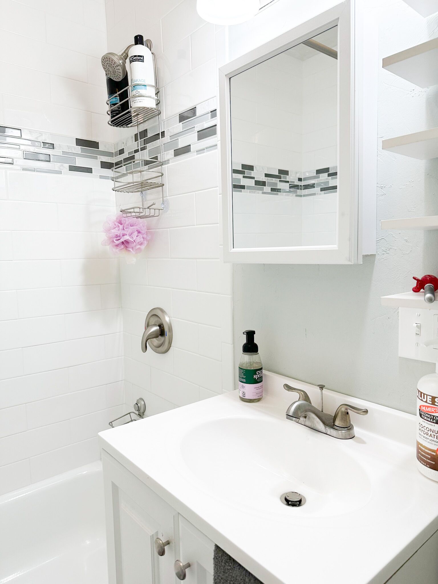 Un bany amb un lavabo blanc, un mirall i una zona de dutxa. Hi ha productes de tocador a la paret i una esponja de bany rosa.