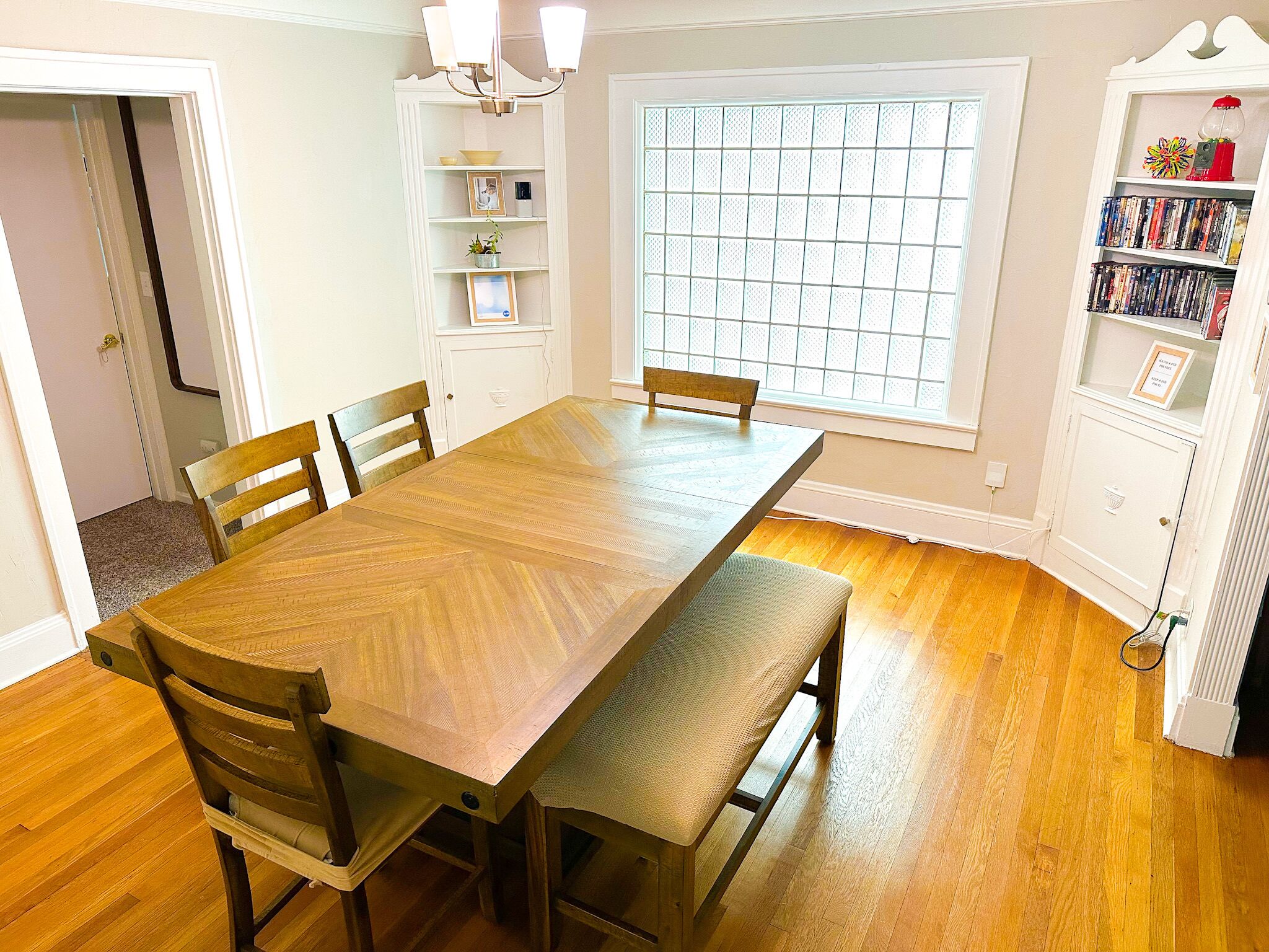 A dining room with a wooden table, four chairs, a bench, a chandelier, a window with blinds, and a bookshelf.