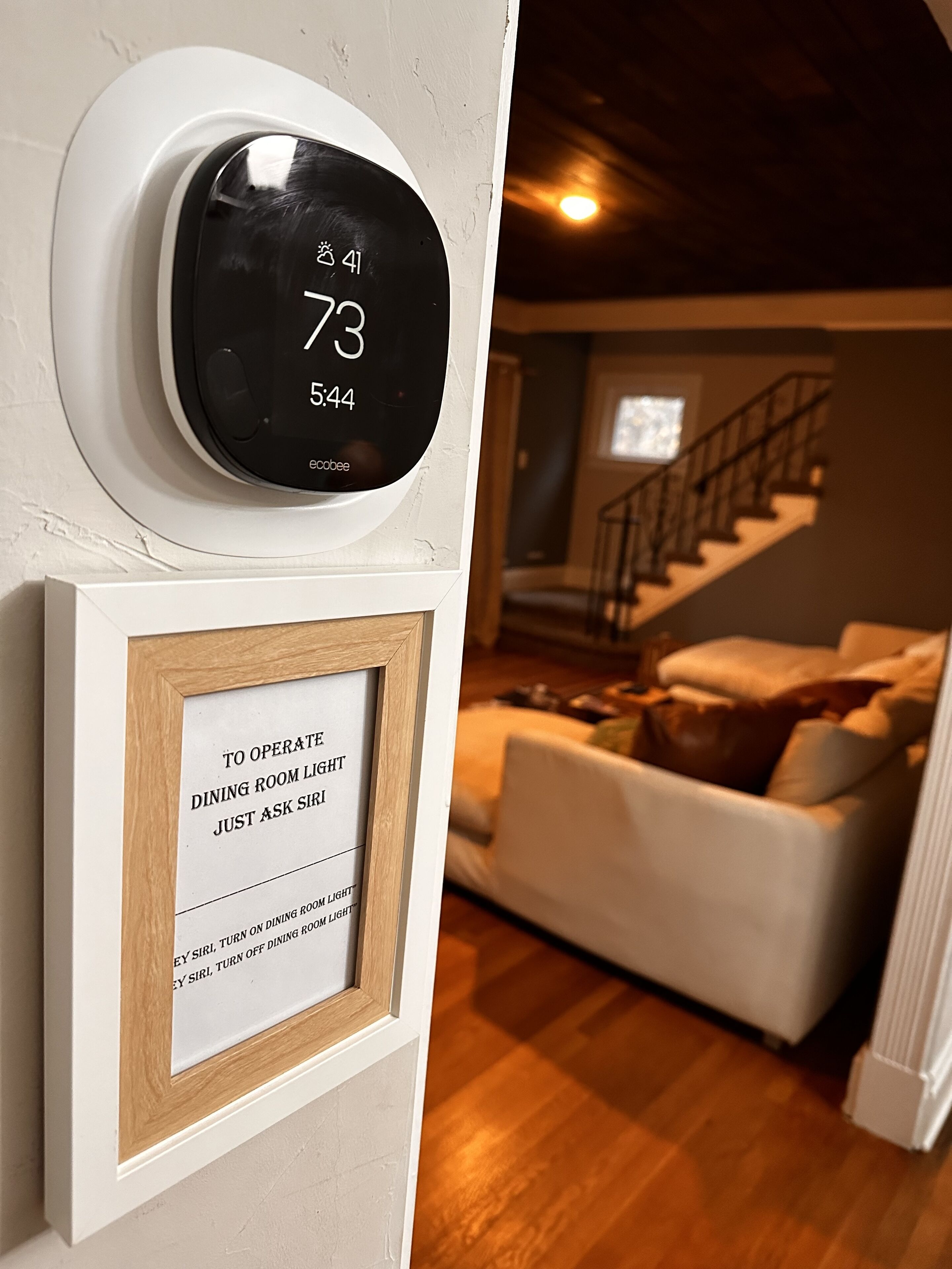 A digital clock on a wall displaying the time as 7:43. Below it, there is a framed sign instructing to ask the maid to turn on the dining room light.