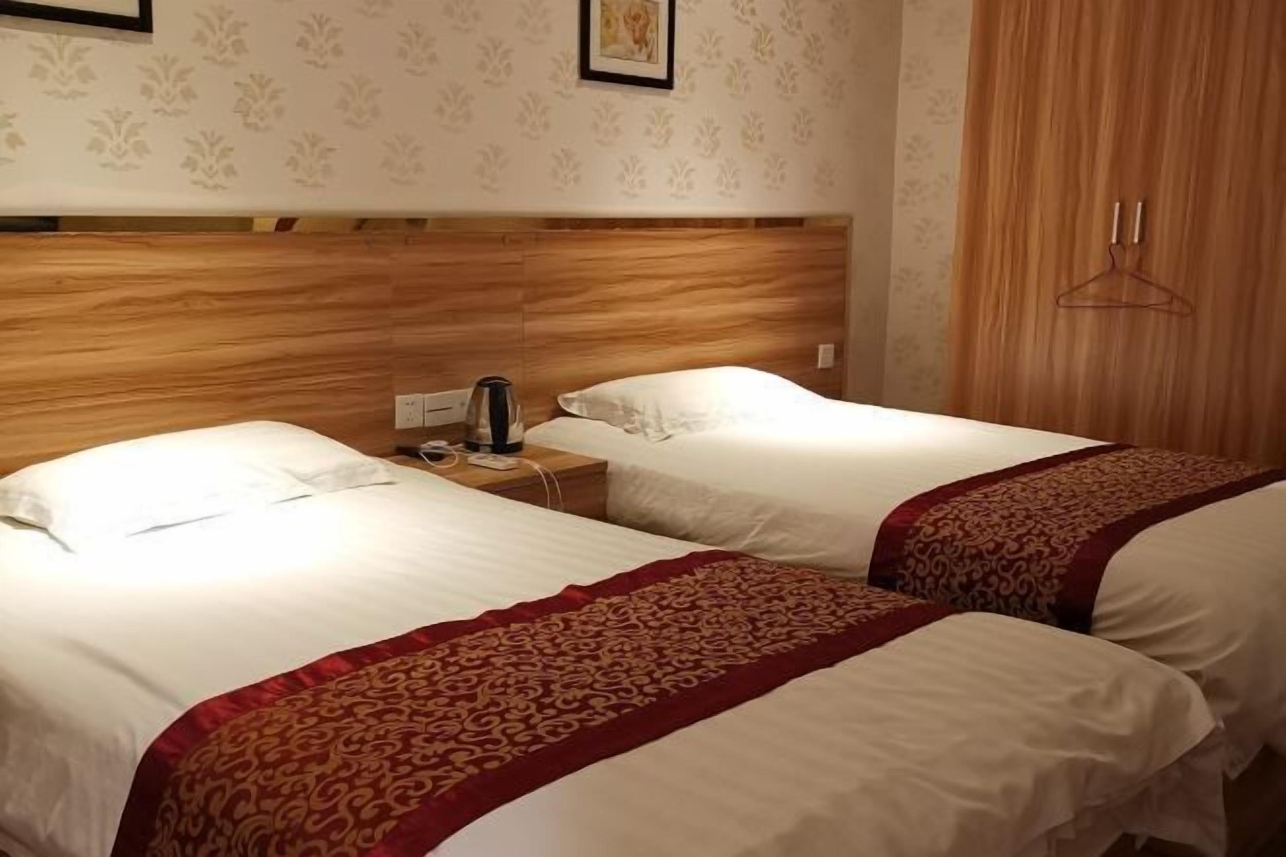 Duas camas de solteiro com lençóis brancos e detalhes vermelhos decorativos, uma cabeceira de madeira, uma mesa de cabeceira com luminária e um cabideiro.