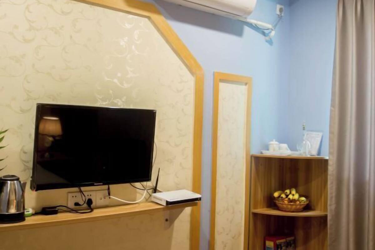 Quarto com televisão sobre suporte de madeira, um dispositivo branco no suporte, ar condicionado fixado na parede e prateleira de madeira com cesta de frutas.