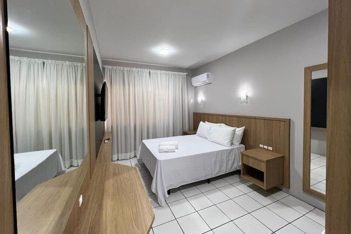 A hotel room with a bed, bedside tables, a mirror, curtains, and an air conditioning unit.