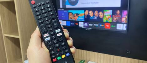 A hand holding a remote control in front of a television displaying a smart TV interface.