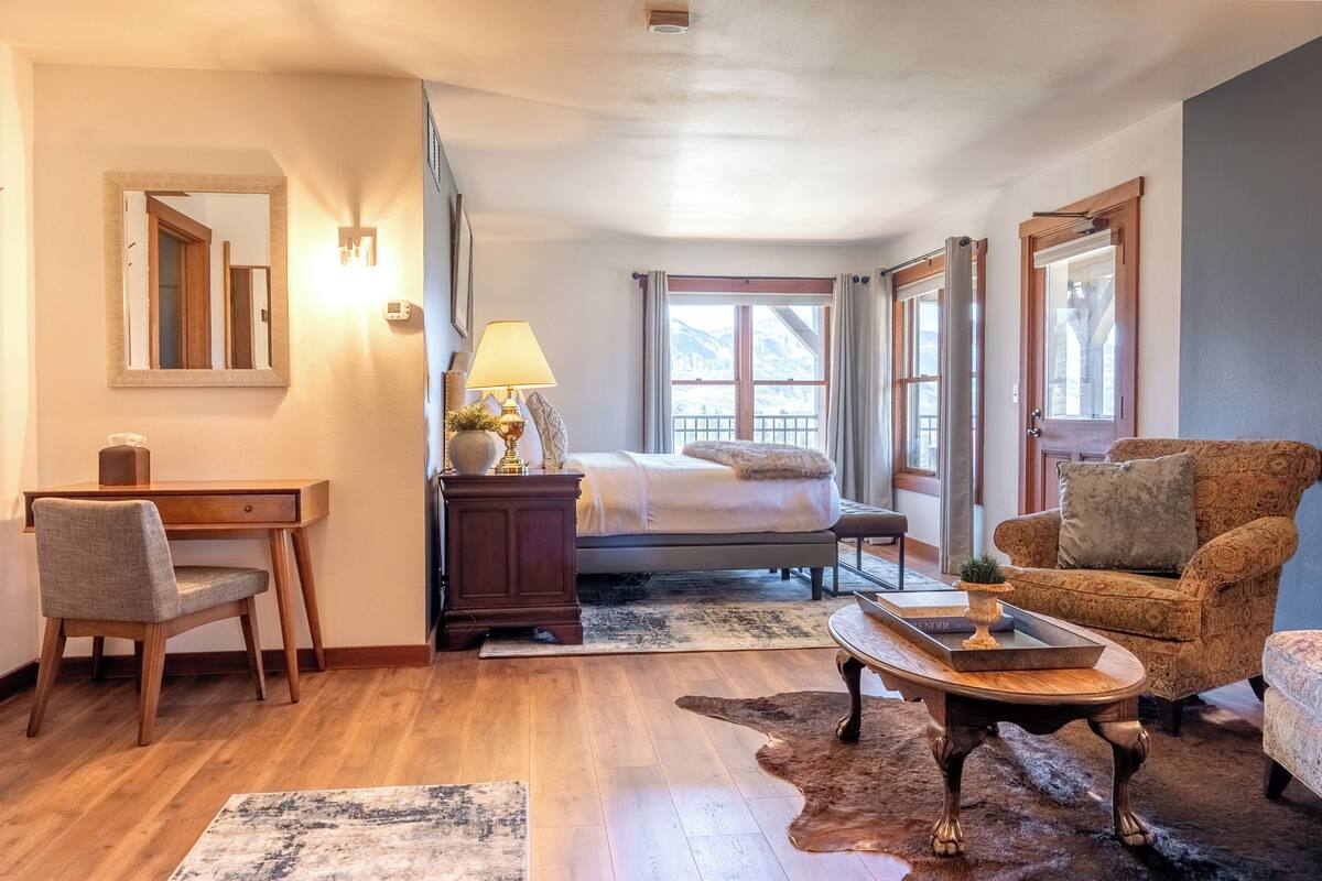 A bedroom with a bed, a desk, a chair, a lamp, a coffee table, and a view of mountains through the window.