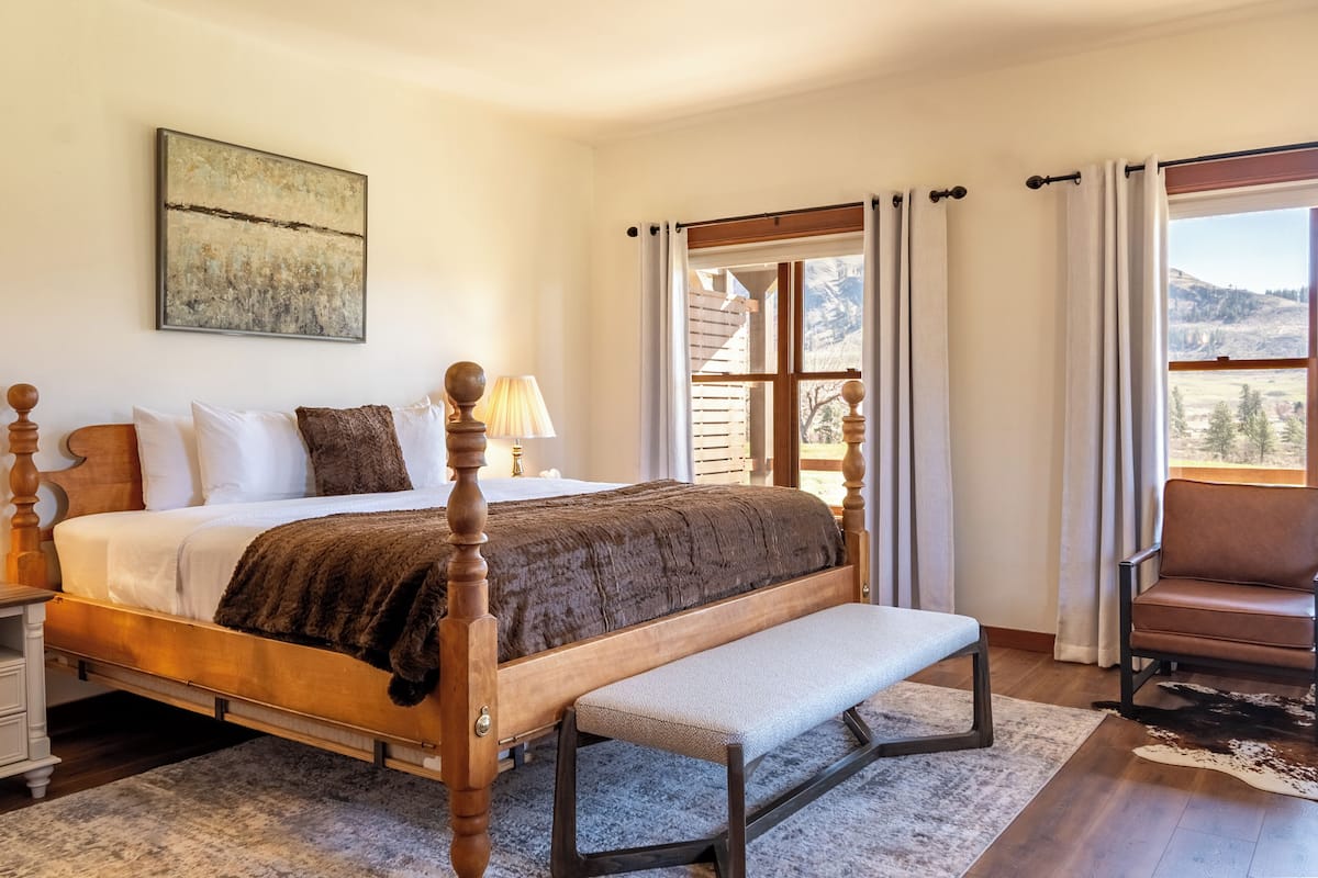 A bedroom with a four-poster bed, a bench, a chair, a window with curtains, and a painting on the wall.