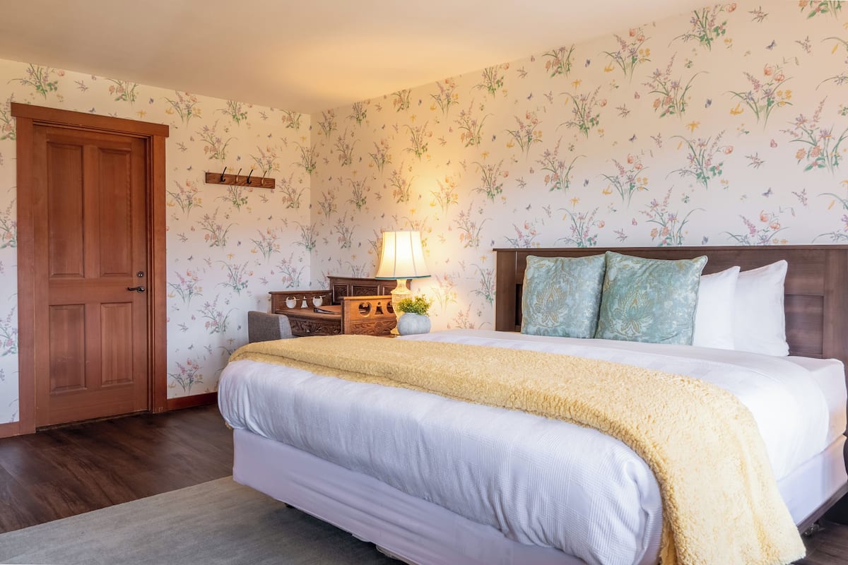 A bedroom with a bed, wooden door, floral wallpaper, and a nightstand with a lamp.