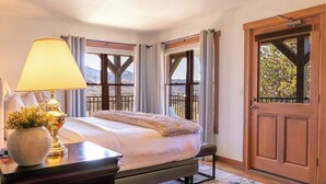 A bedroom with a bed, a lamp, a vase with flowers, a wooden door, and a view of mountains.