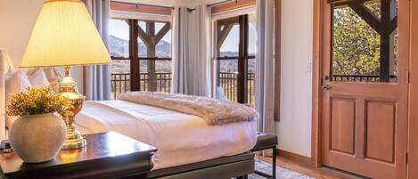 Un dormitorio con cama, lámpara, un jarrón con flores, puerta de madera y vistas a las montañas.