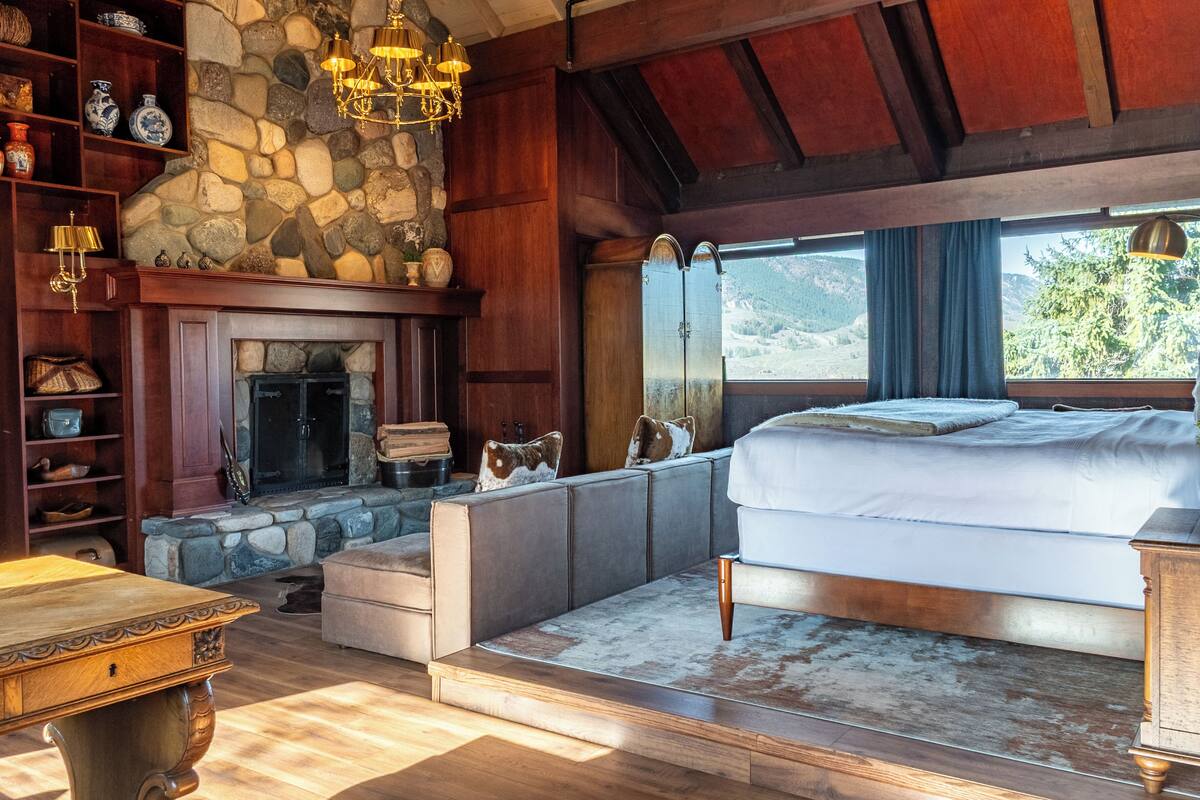 A bedroom with a stone fireplace, a large bed, a wooden ceiling, and a view of the outdoors.