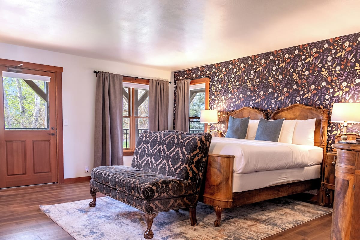 A bedroom with a bed, a patterned armchair, a wooden nightstand, and a floral wallpaper.