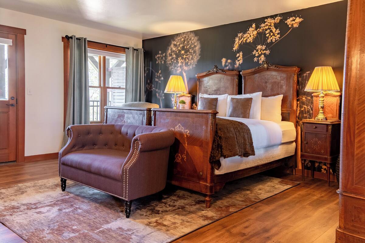 A bedroom with a wooden bed, two bedside tables with lamps, a brown armchair, and a large window with curtains.