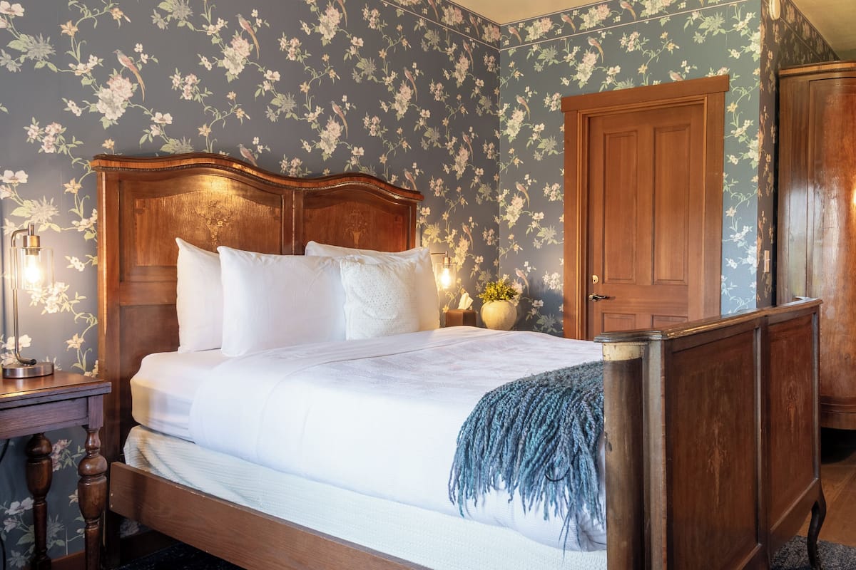A bedroom with a wooden bed, floral wallpaper, a nightstand with a lamp, and a wooden wardrobe.
