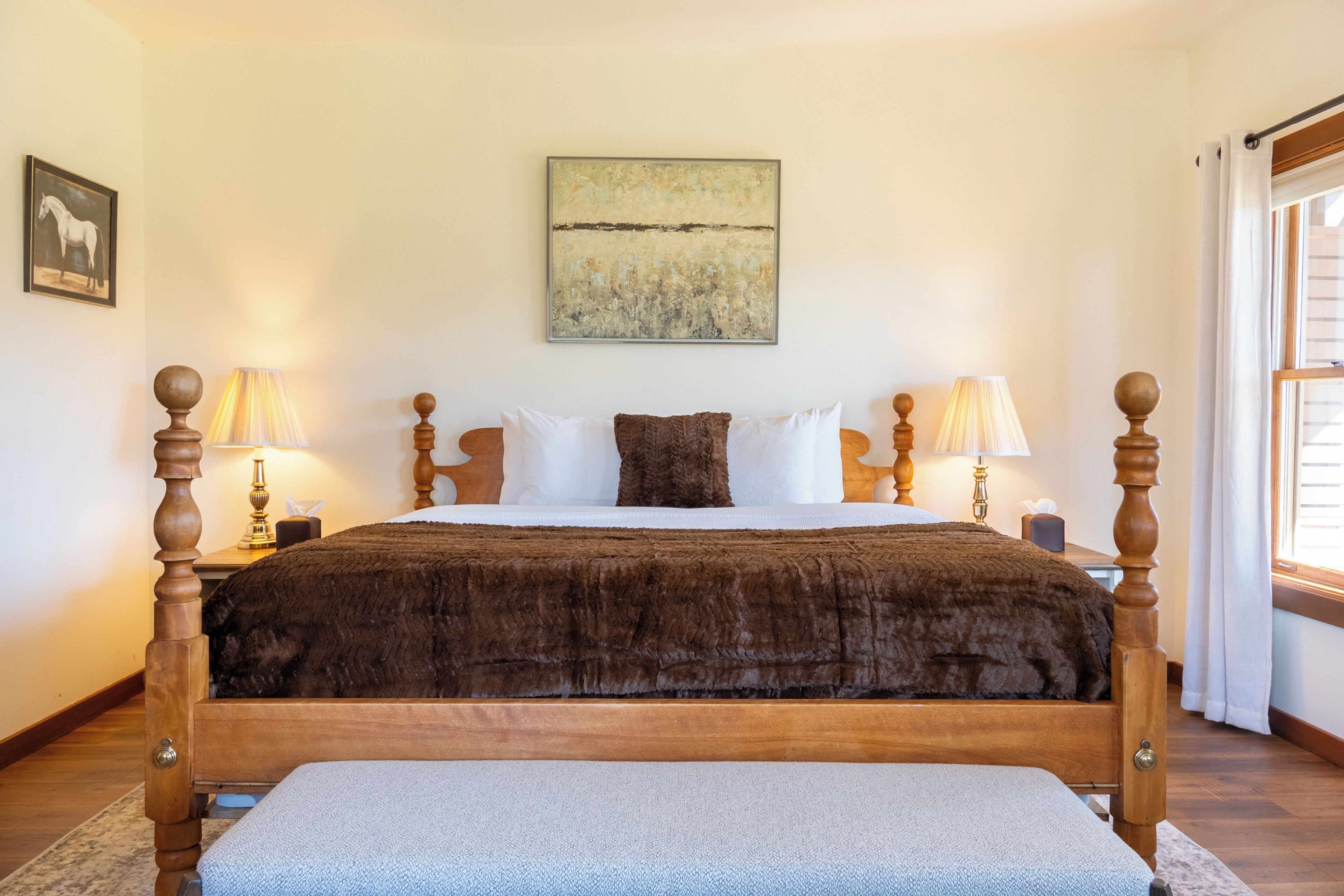 A neatly made bed with a wooden frame and a plush brown blanket, flanked by two bedside lamps, a framed picture on the wall, and a window with curtains.