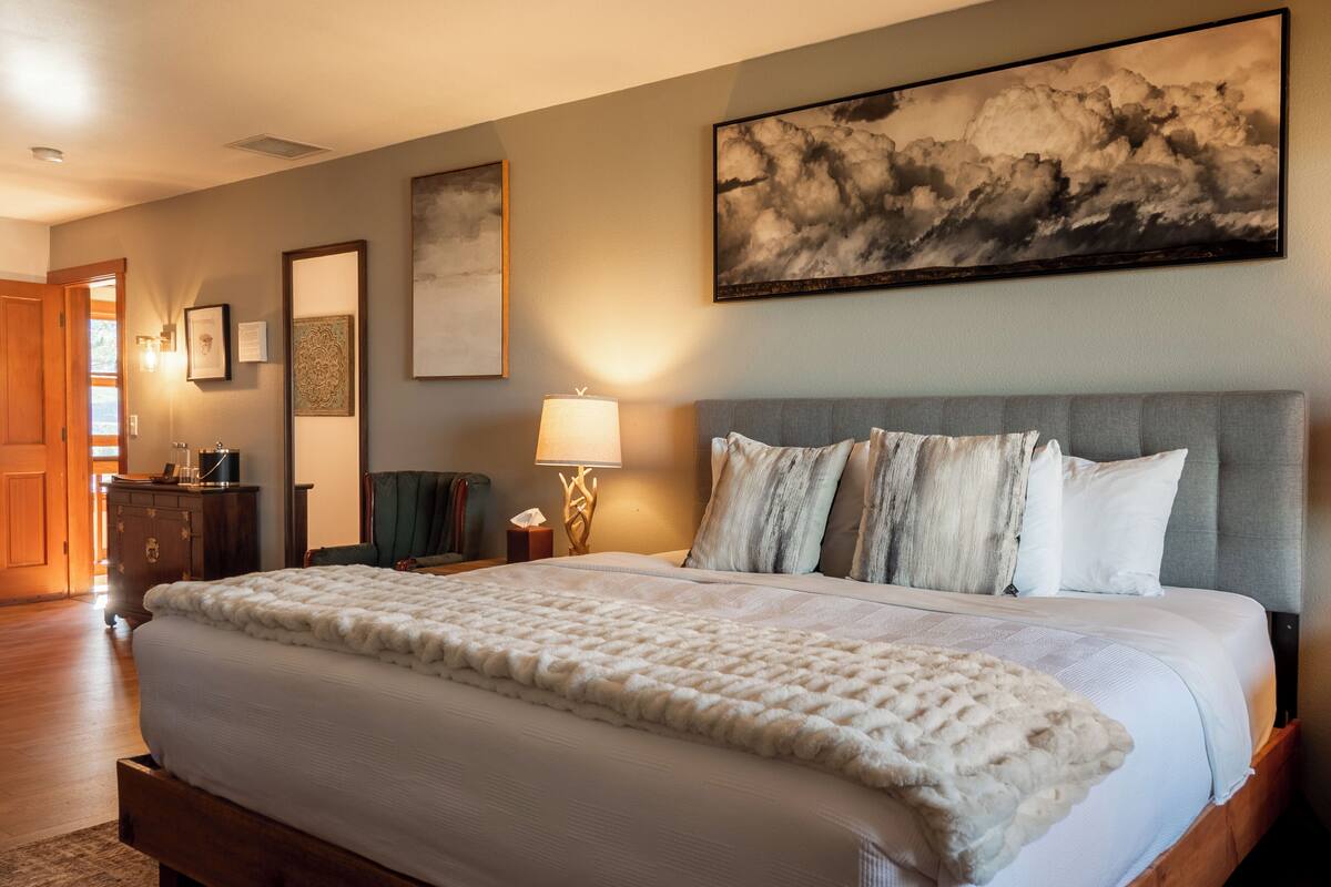 A bedroom with a large bed, a headboard, a nightstand with a lamp, and a wall-mounted artwork.