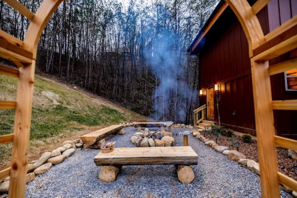 A rustic outdoor seating area with a fire pit, surrounded by wooden logs and rocks, leading to a wooden structure with a chimney emitting smoke.