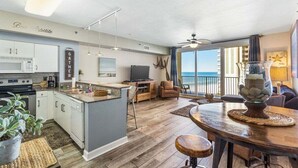 A modern kitchen with a central island, a dining area with a wooden table, and a balcony with a view of the ocean. - Shores of Panama 926- Free Beach Chairs! (Panama City Beach)