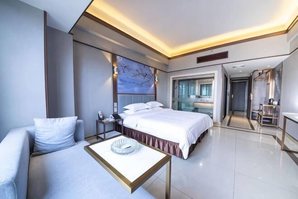 Habitación de hotel moderna con cama, sofá, mesita de noche, espejo y vista al baño.