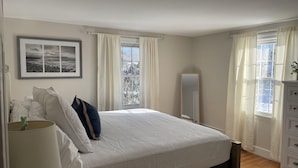 A bedroom with a bed, a bedside lamp, a mirror, and a window with curtains. - Adorable, Bright and By the Bay! (Falmouth)