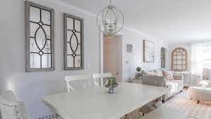 A dining area with a white table and chairs, a chandelier, and a view of a living room with a sofa and a window. - Adorable, Bright and By the Bay! (Falmouth)