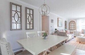 Uma sala de jantar com uma mesa e cadeiras brancas, um candelabro e vista para uma sala de estar com um sofá e uma janela.