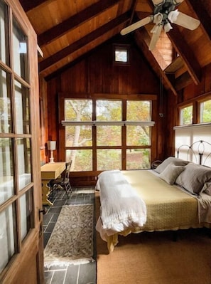 Cozy bedroom with a sloped ceiling and wooden beams, featuring a large bed and a desk with a chair near a window offering a view of the outdoors.
