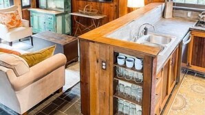 Ein rustikales Blockhaus-Interieur mit Holzbalkendecke, einer zentralen Kücheninsel mit Spüle, einem an der Wand befestigten Fernseher und einer gemütlichen Sitzecke mit Sofa und Sesseln.