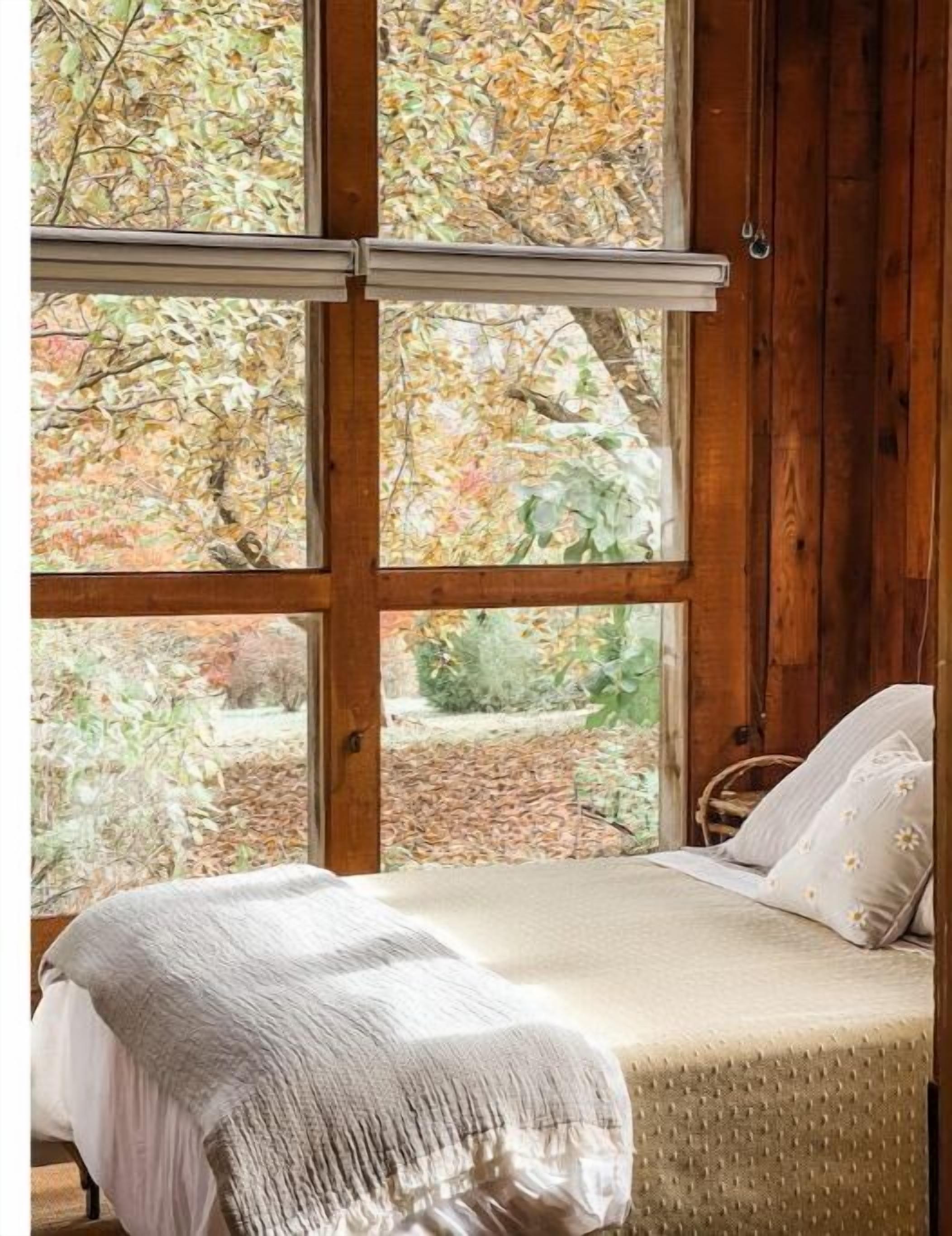 Ein Schlafzimmer mit großem Fenster, einem Bett mit weißer Bettdecke und Blick auf Bäume und Grünpflanzen.